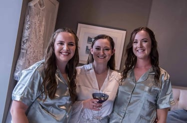 Smiling bride in a white lace robe and bridesmaids in sage green silk pajamas getting ready.
