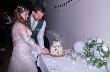 A bride and groom smiling while cutting their two-tier white wedding cake decorated with pressed flowers.