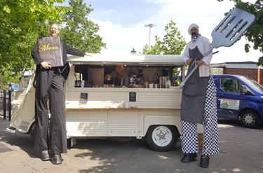 Performers on stilts dressed as a waiter and chef posing by a vintage mobile catering food truck.