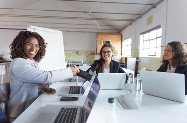 Professional business women in a modern office shaking hands over laptops during a successful meeting.