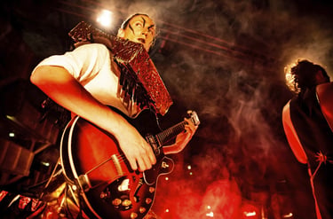 A theatrical guitarist with face paint plays a hollow body electric guitar on a smoky stage.