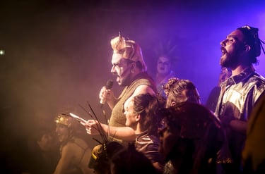 A drag performer in gold sequins sings into a microphone during a vibrant live theater production.
