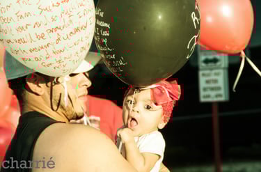 baby with red hair bow being held by father in the middle of a sea of balloons