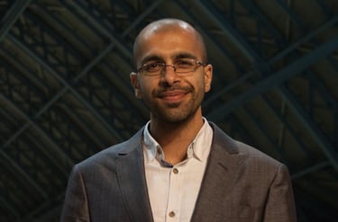 A smiling professional man with glasses and a grey blazer in a modern office setting.
