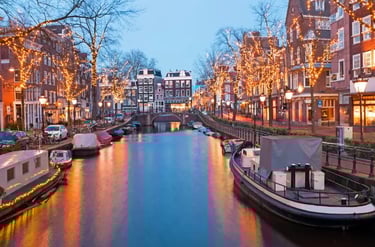 Amsterdam canal at dusk with festive Christmas lights on trees and historic Dutch architecture reflecting on the water.