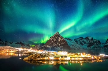 Vibrant green Northern Lights aurora borealis over a snowy mountain and lit village in Reine, Lofoten Islands.