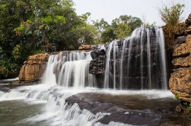 Long exposure scenic waterfall flowing over dark layered rocks surrounded by lush green tropical trees.
