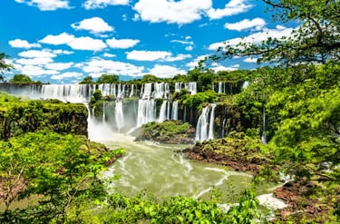 Panoramic view of Iguazu Falls with cascading waterfalls and lush green tropical rainforest under a blue sky.