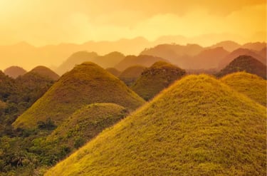 Golden Chocolate Hills of Bohol, Philippines, under a warm sunset sky with misty mountains.