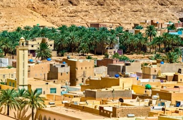Scenic view of a traditional desert town in Algeria featuring a tall minaret, palm trees, and tan buildings.
