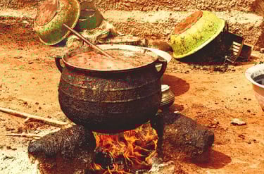 A large black cast iron pot simmering with stew over an open wood fire in a rural outdoor kitchen.