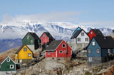 Colorful coastal houses in a Greenlandic village with snowy mountain peaks and clouds in the background.