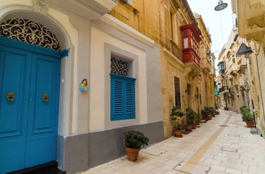 A narrow limestone street in Malta featuring traditional blue doors and red wooden balconies.