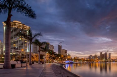 Sunset over Luanda Bay skyline with illuminated buildings and palm trees along the waterfront promenade.