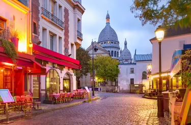 A charming cobblestone street in Montmartre, Paris, featuring local cafes and the Sacré-Cœur Basilica at dusk.