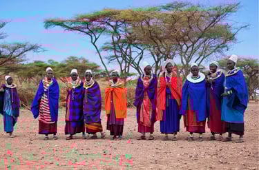 Maasai women in traditional colorful robes and beaded jewelry stand in a Kenyan savanna landscape.