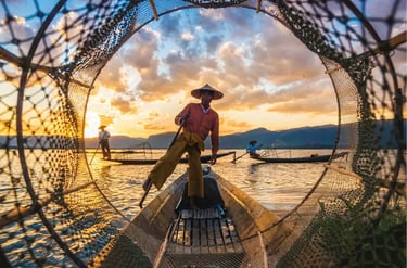 Intha fisherman rowing a wooden boat on Inle Lake at sunset using traditional leg-rowing technique.