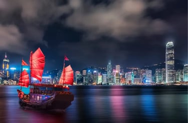 A traditional red-sailed junk boat cruises Victoria Harbour against the illuminated Hong Kong skyline at night.