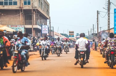 Busy street in Benin with people riding motorcycles and scooters past local market shops.