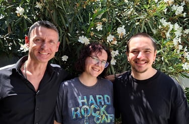 Three smiling friends posing together outdoors in front of a blooming white oleander bush.