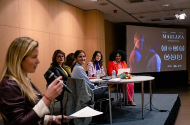 A panel of female speakers sits at a table during a film presentation for Mariana in an auditorium.