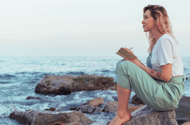 Woman by the ocean holding a journal and pen, reflecting on emotional intelligence & self-awareness.