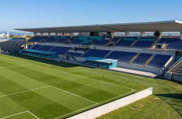 Empty outdoor football stadium with blue seats and green grass field under a clear blue sky.