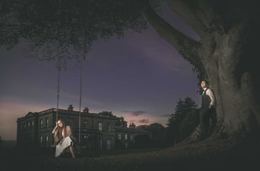 A couple poses for a romantic wedding portrait by a large tree and swing outside a historic manor at twilight.