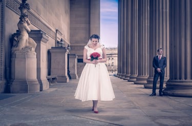 A bride in a vintage tea-length wedding dress holds red roses outside a neoclassical building with columns.