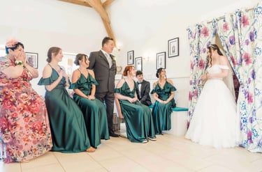 A group of bridesmaids in emerald green gowns and family members reacting to a bride in her wedding dress.