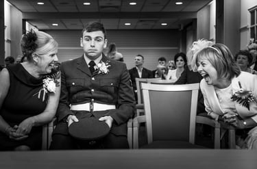 A young groom in a military dress uniform sits between two women laughing at a wedding ceremony.