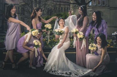 Smiling bride in a white lace gown surrounded by bridesmaids in lavender dresses holding white rose bouquets.