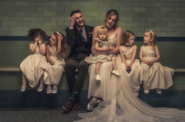 A bride and groom in wedding attire sitting with five flower girls in white dresses against a green tiled wall.