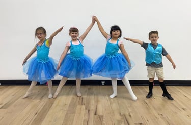 Young children in blue ballet tutus and dance costumes posing in a dance studio with wood floors.