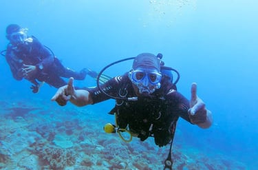 Two scuba divers wearing full gear explore a vibrant coral reef in clear blue ocean water.