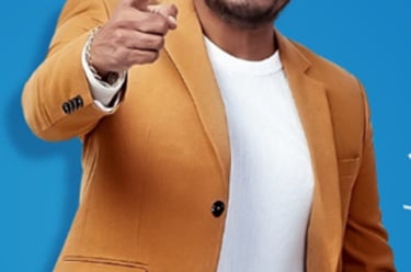 Kannada actor Shiva Rajkumar pointing forward while wearing a stylish mustard yellow blazer over a white shirt.