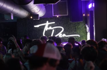 Crowded nightclub dance floor with a neon 'The Den' sign on a green foliage wall.