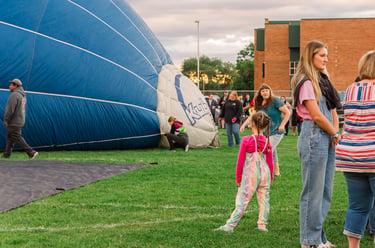 Spectators watch a large blue hot air balloon being inflated on a green field during a community festival.
