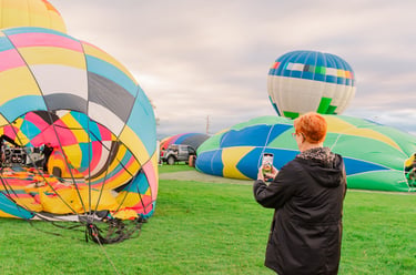 A woman takes a photo of colorful hot air balloons inflating on a green field during a festival.