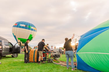 A flight crew prepares colorful hot air balloons for takeoff in a grassy field during a festival.