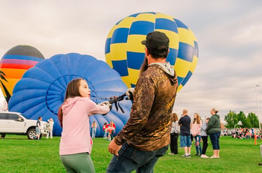 A young girl and man pull ropes to inflate colorful hot air balloons at an outdoor festival.