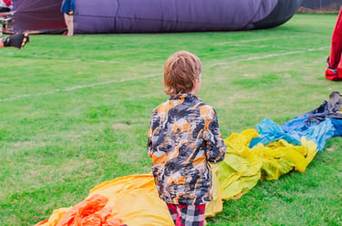 A young boy watches a colorful rainbow hot air balloon being inflated on a green grass field.