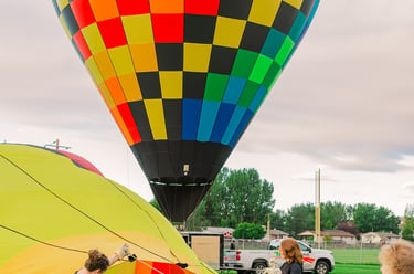 A vibrant rainbow checkered hot air balloon inflates on a grassy field with a ground crew.