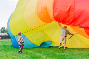 A child watches as a man prepares a colorful rainbow hot air balloon for inflation on a grassy field.