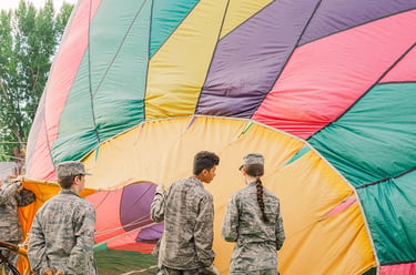 Military cadets in camouflage uniforms preparing a colorful hot air balloon for flight on a grassy field.