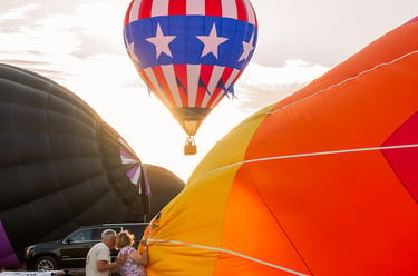 A vibrant patriotic hot air balloon takes flight as crews inflate more balloons during a sunset festival.
