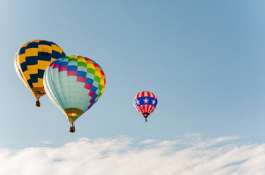 Three colorful hot air balloons floating in a clear blue sky with light clouds during a festival.