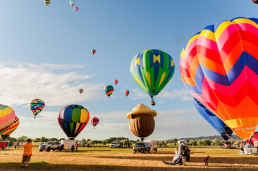 Colorful hot air balloons taking off from a field during a morning festival under a clear blue sky.