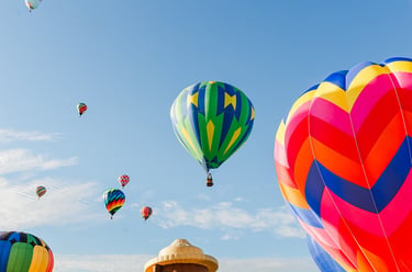 Colorful hot air balloons in various shapes and sizes take flight over a grassy field during a festival.