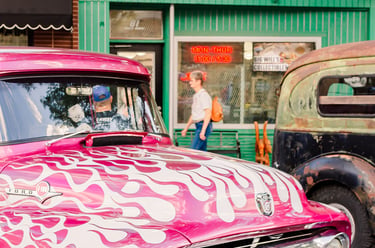 Vintage pink Ford F100 truck with white flame decals parked on a city street near storefronts.